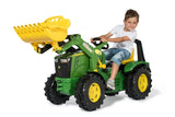Child riding a green Rolly X Trac John Deere toy tractor with a yellow loader bucket on a white background