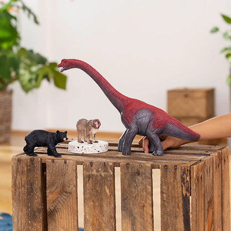 Schleich American Black Bear figure beside a red long neck dinosaur and a small grey animal figure on a wooden crate.