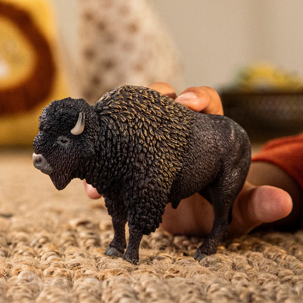 Schleich Bison figure standing on a woven carpet while a hand holds its back, showing the thick textured coat and short curved horns