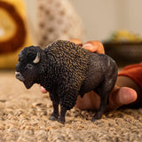 Schleich Bison figure standing on a woven carpet while a hand holds its back, showing the thick textured coat and short curved horns