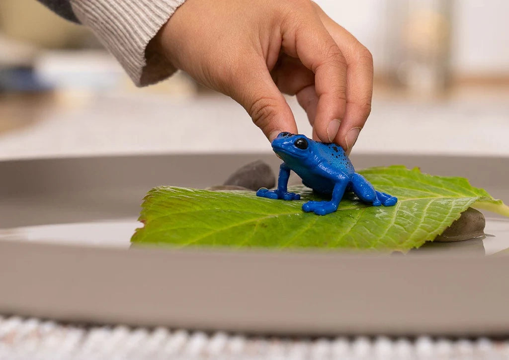 Schleich Blue Poison Dart Frog figure standing on a green leaf as a hand reaches down from above.