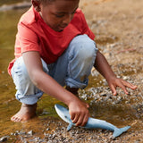 Child holding the Schleich Blue Whale figure on a rocky shoreline beside shallow water