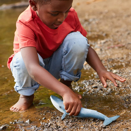 Child holding the Schleich Blue Whale figure on a rocky shoreline beside shallow water