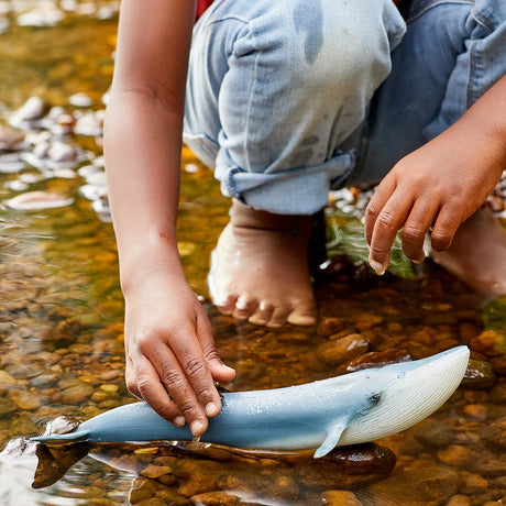 Schleich Blue Whale figure in shallow water beside a child’s hand and bare feet