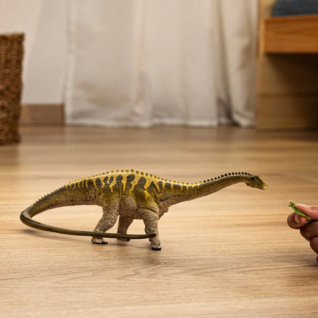 Schleich Diplodocus figure on a wooden floor beside a hand holding a small green object