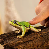 A green tree frog figurine on a piece of wood, shown with a hand beside it for scale and its side stripe clearly visible.