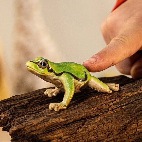 A green tree frog figurine on a piece of wood, shown with a hand beside it for scale and its side stripe clearly visible.