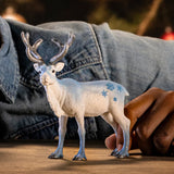 Schleich Frosty Christmas Reindeer figure standing on a table beside a hand, with a dark blue shirt in the background. 