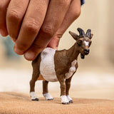 Schleich Goat figure standing on tan fabric while a hand rests on its back, showing curved horns and a white forehead marking