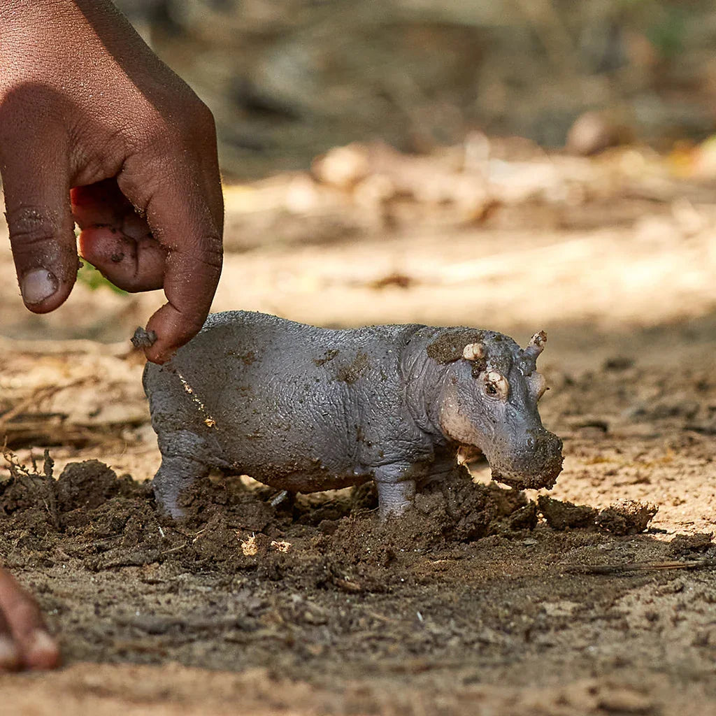 Schleich hippopotamus figure partly covered in soil with a hand beside it.
