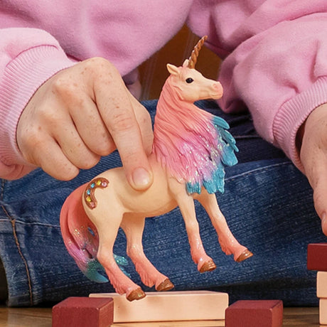 Hands holding the Schleich Marshmallow Unicorn Mare figure above wooden blocks, showing its colourful mane, tail and horseshoe marking