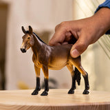Schleich Mule figure standing on a wooden surface while a hand holds its back.