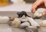 Schleich Otter figure standing among smooth rocks while a hand holds a stone above the rocks.