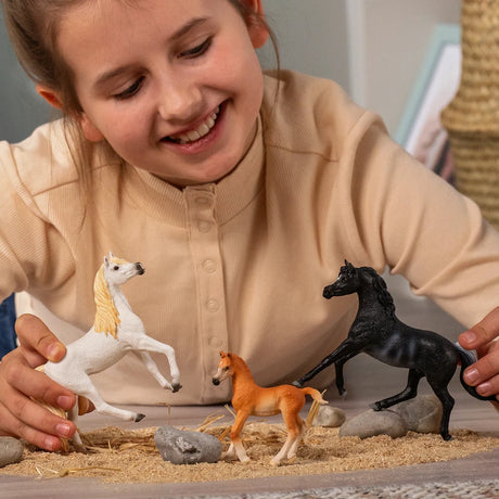 Schleich Rabicano Arabian Stallion figure beside a white horse and chestnut foal as hands arrange the figures among sand, rocks and straw.