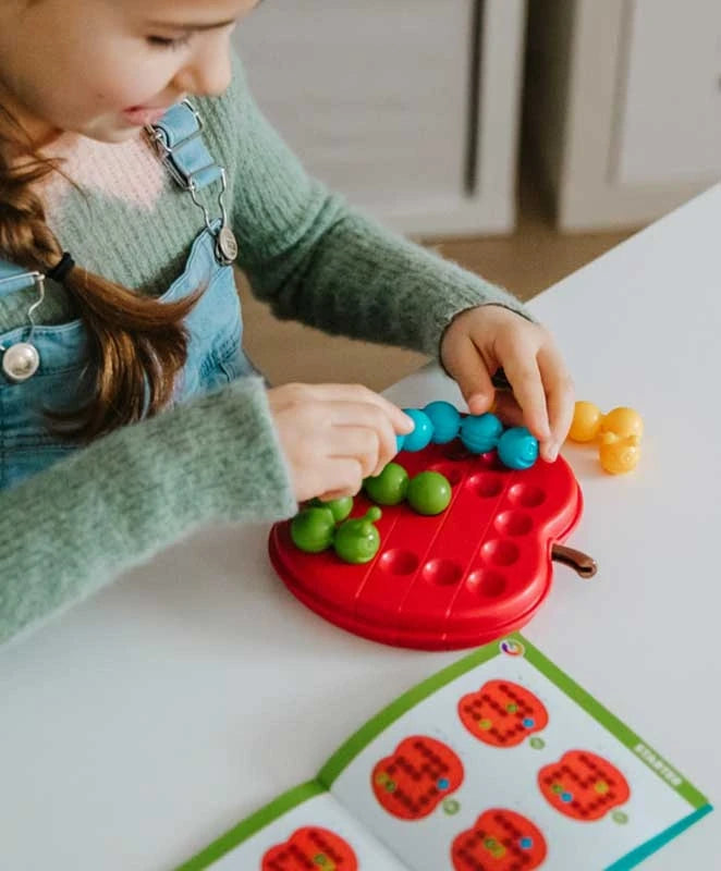 Hands placing blue and green caterpillar pieces on a red apple-shaped board beside an open challenge booklet