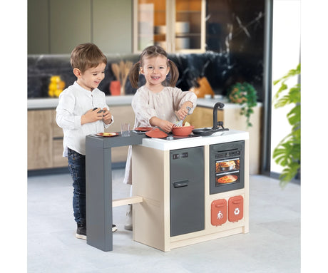 Two children playing with the Smoby Kitchen Island in a home kitchen, with a girl stirring a cooking pot and a boy standing beside the dining counter.