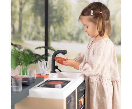 Girl holding an orange toy plate beside the Smoby Kitchen Island sink, with black tap, cooking hob, frying pan and kitchen utensils visible.