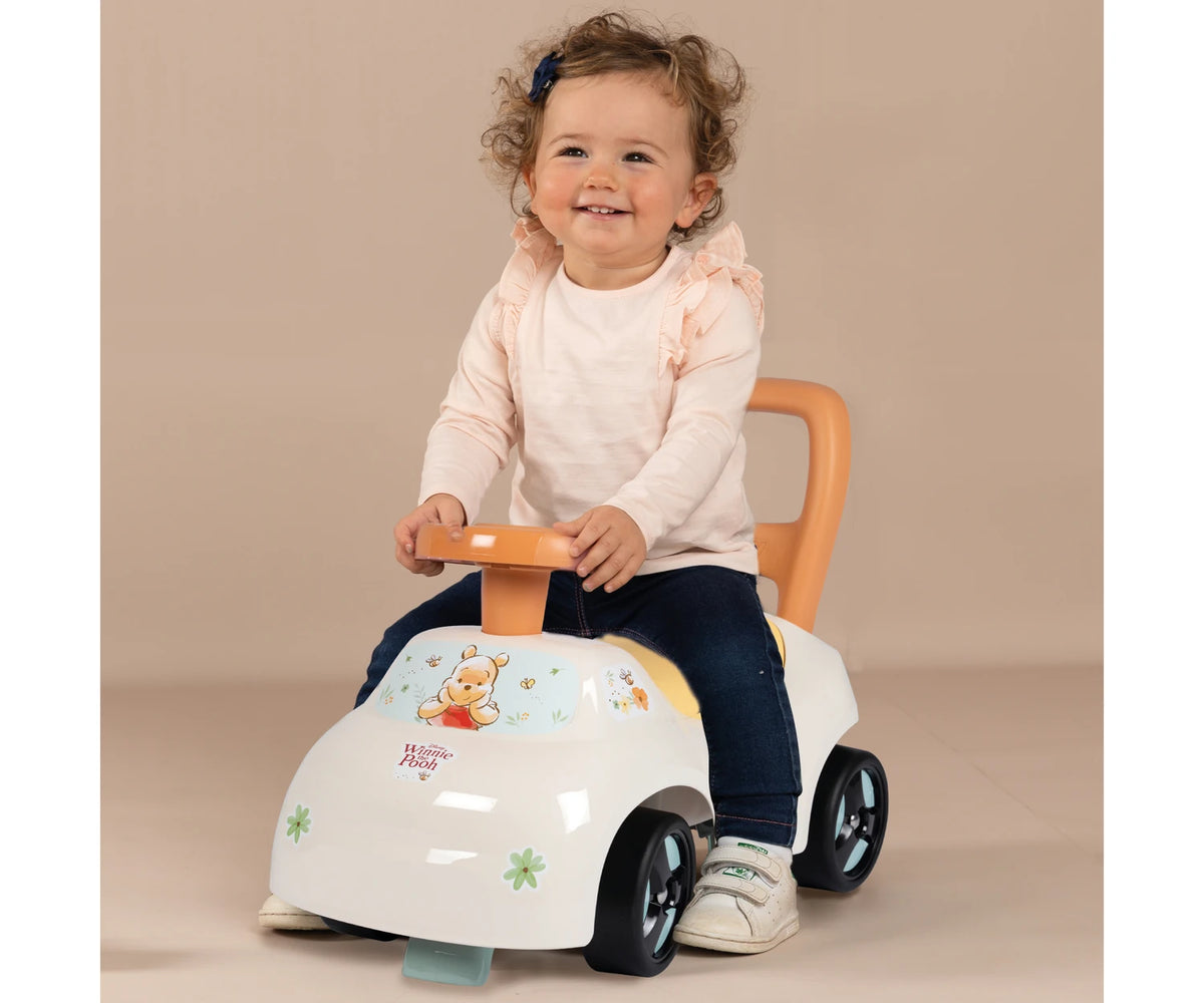 Young child sitting on the white Smoby Winnie the Pooh ride on car, holding the orange steering wheel with Winnie the Pooh illustrations visible on the front.