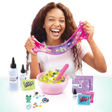 Smiling girl stretching pink and purple slime filled with colourful decorations, with a pink mixing bowl, ingredient bottles, spatula, slime sachets and small charms displayed on the table.