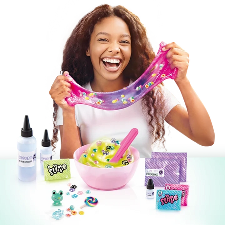 Smiling girl stretching pink and purple slime filled with colourful decorations, with a pink mixing bowl, ingredient bottles, spatula, slime sachets and small charms displayed on the table.