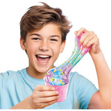 Smiling boy wearing a light blue T-shirt stretching a long strand of colourful transparent slime from a pink pot, with miniature fruit decorations mixed throughout.