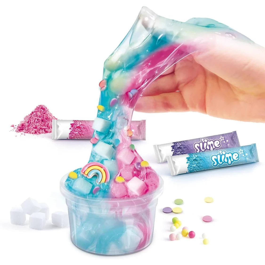 A hand stretching transparent pink and blue So Slime from a clear pot, decorated with a miniature rainbow, white cubes and colourful pieces, with three coloured dye sachets beside it.