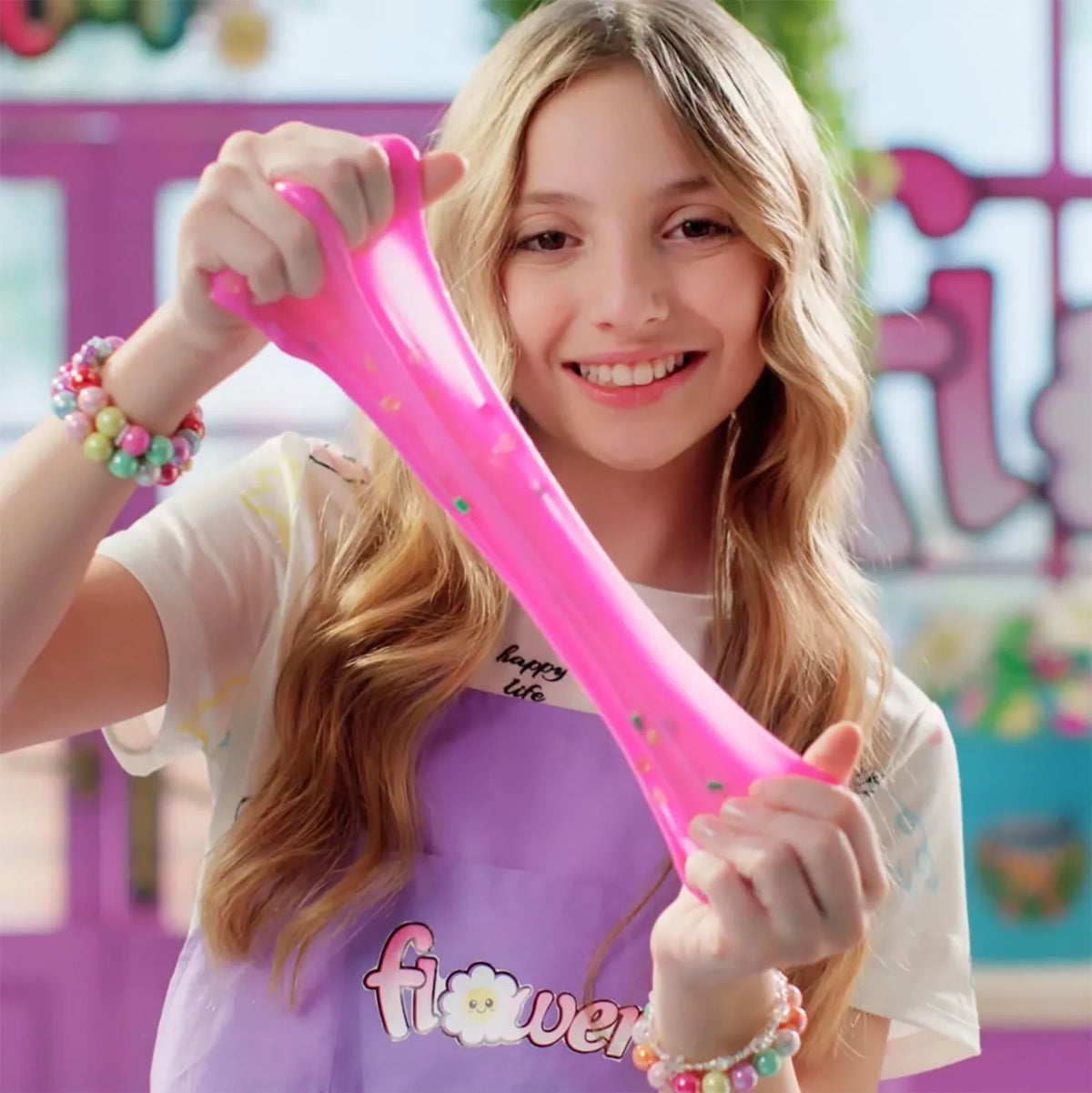 Smiling girl wearing a purple Flower Slime apron and colourful bead bracelets, stretching a long strand of bright pink slime containing small decorative pieces between both hands.