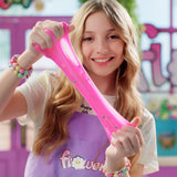 Smiling girl wearing a purple Flower Slime apron and colourful bead bracelets, stretching a long strand of bright pink slime containing small decorative pieces between both hands.