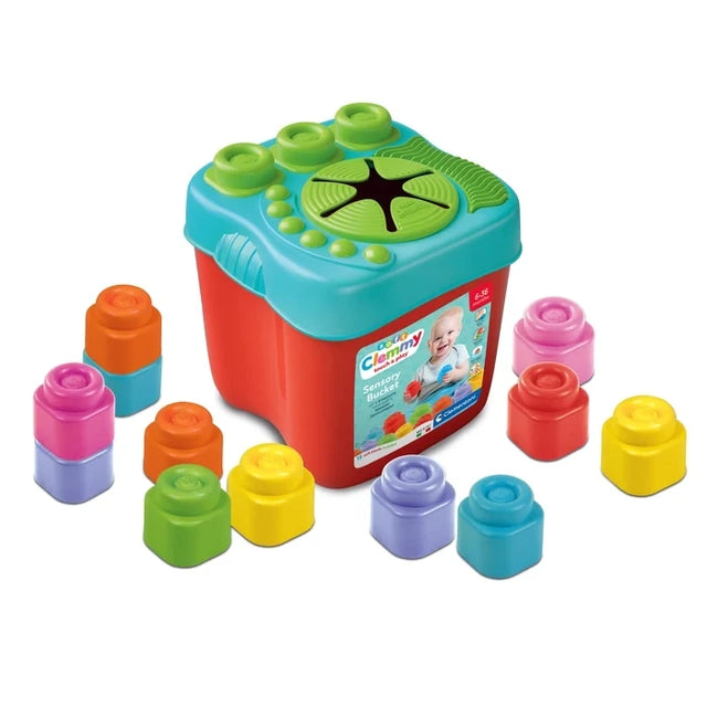 Soft Clemmy Sensory Bucket with red storage container, turquoise textured lid with green stacking studs and flower shaped opening, surrounded by colourful soft building blocks.