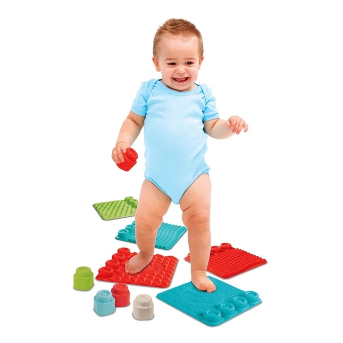 Smiling baby wearing a light blue bodysuit standing barefoot on red and turquoise Soft Clemmy sensory tiles, with additional textured tiles and colourful blocks on the floor.