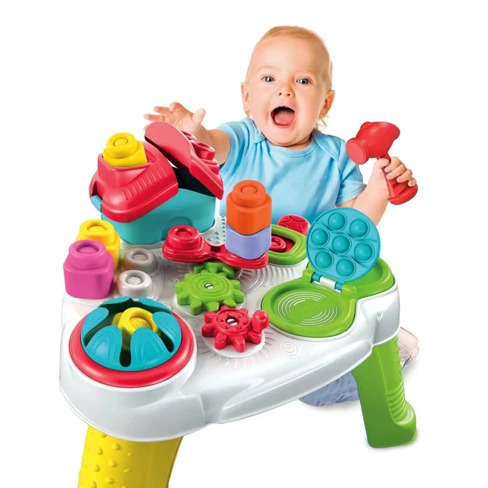 Smiling baby in a blue shirt holding a red toy hammer behind the Soft Clemmy Sensory Table, with colourful stacking blocks, red and green gears and blue pop-it panel.