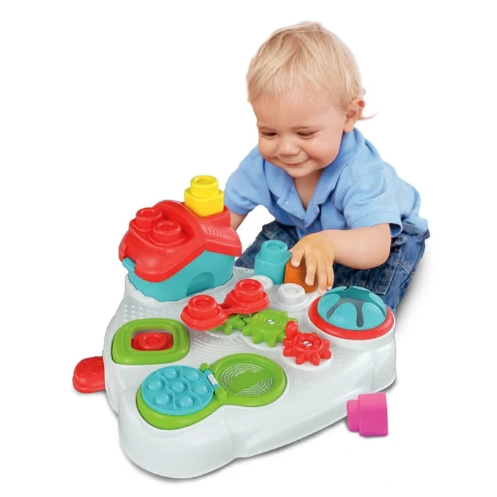 Young child in a blue shirt sitting on the floor and reaching towards the Soft Clemmy Sensory Table, showing its red roofed house, colourful gears and textured activity panels.