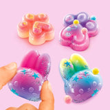 Four decorated Squish Squish squishies on a pink background, featuring two glittery paw shapes and two bunny faces in pink, purple, blue and yellow, with star confetti and small pom poms.