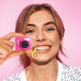 Smiling woman holding the small pink Style 4 Ever digital camera beside her face, showing the front camera lens, flash and attached yellow keychain clip against a pink background.