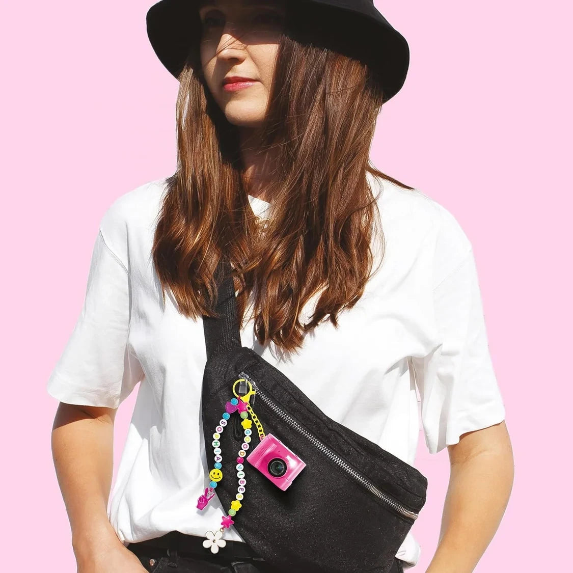 Woman wearing a white T-shirt and black hat with a black crossbody bag decorated with the pink Style 4 Ever Mini Cam, colourful alphabet beads, a yellow clip and dangling flower and hand charms.