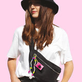 Woman wearing a white T-shirt and black hat with a black crossbody bag decorated with the pink Style 4 Ever Mini Cam, colourful alphabet beads, a yellow clip and dangling flower and hand charms.