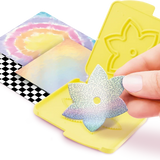 Hand holding an iridescent paper flower cutout above an open yellow flower cutting plate, with pastel coloured paper sheets and black and white checked paper underneath.