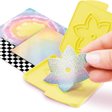 Hand holding an iridescent paper flower cutout above an open yellow flower cutting plate, with pastel coloured paper sheets and black and white checked paper underneath.