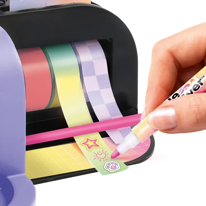 Close-up of a hand using a pink marker to decorate adhesive tape printed with stars, a sun and paw print, with pink, green and purple tape rolls mounted inside the Style 4 Ever Scrapbooking Studio Pro.