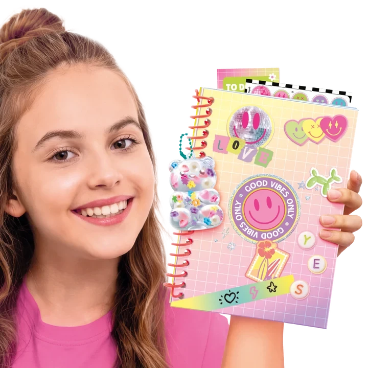 Smiling girl holding a pink spiral bound scrapbook decorated with smiley faces, colourful hearts, stickers, a transparent bear decoration, the word LOVE and a circular Good Vibes Only sticker.