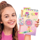 Smiling girl holding a pink spiral bound scrapbook decorated with smiley faces, colourful hearts, stickers, a transparent bear decoration, the word LOVE and a circular Good Vibes Only sticker.