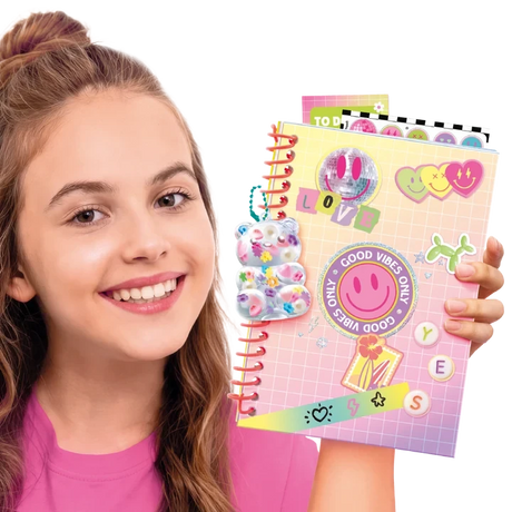 Smiling girl holding a pink spiral bound scrapbook decorated with smiley faces, colourful hearts, stickers, a transparent bear decoration, the word LOVE and a circular Good Vibes Only sticker.