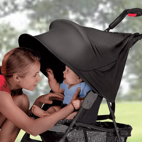 Black stroller shade fitted to a stroller, with a woman leaning in toward a baby seated under the extended canopy, showing the stroller handle with a red button, grey frame parts, and the shade fabric pulled forward