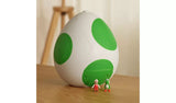 Closed white Yoshi egg with three large green spots standing on a wooden table, with small Fire Mario and Yoshi figures positioned in front.