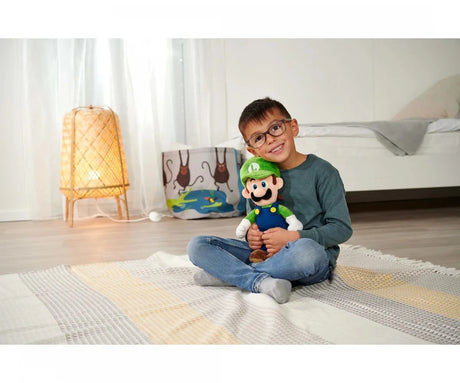 Child sitting cross-legged on a rug holding a Super Mario Luigi plush toy up to their face, showing the toy's size relative to a school-age child