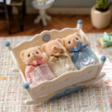 Sylvanian Families Biscuit Bear Sleepy Triplets resting together in their white rocking cradle on a patterned rug, with the three babies dressed in pink, yellow and blue lace gowns.