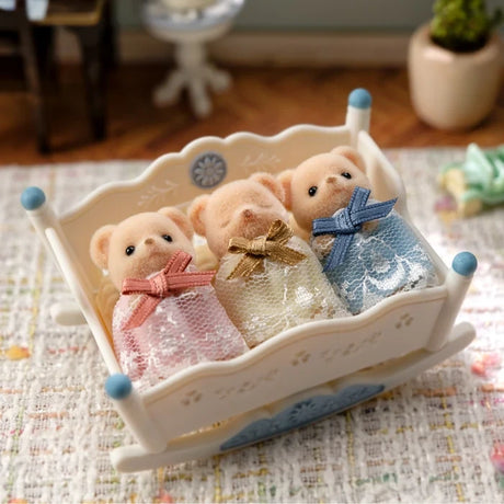 Sylvanian Families Biscuit Bear Sleepy Triplets resting together in their white rocking cradle on a patterned rug, with the three babies dressed in pink, yellow and blue lace gowns.