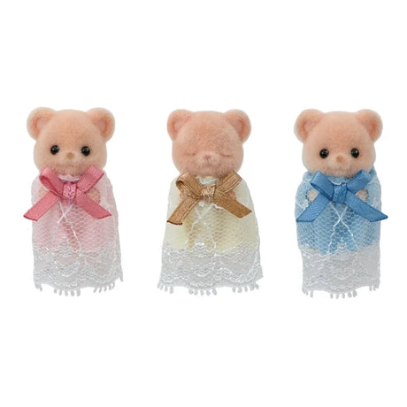Three Sylvanian Families Biscuit Bear baby figures standing separately against a white background, wearing pink, yellow and blue lace gowns with large matching bows. The middle baby has closed eyes.