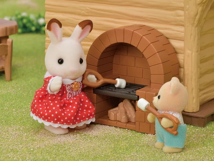 Sylvanian Families rabbit figures roasting marshmallows on sticks beside the Lakeside Lodge brick stove, including a white rabbit wearing a red dress.
