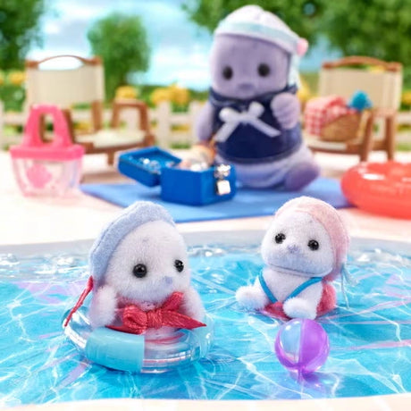 Two Sylvanian Families baby seals playing in blue swimming pool water, one sitting in a transparent blue swimming ring and the other beside a purple beach ball, with their father in the background.